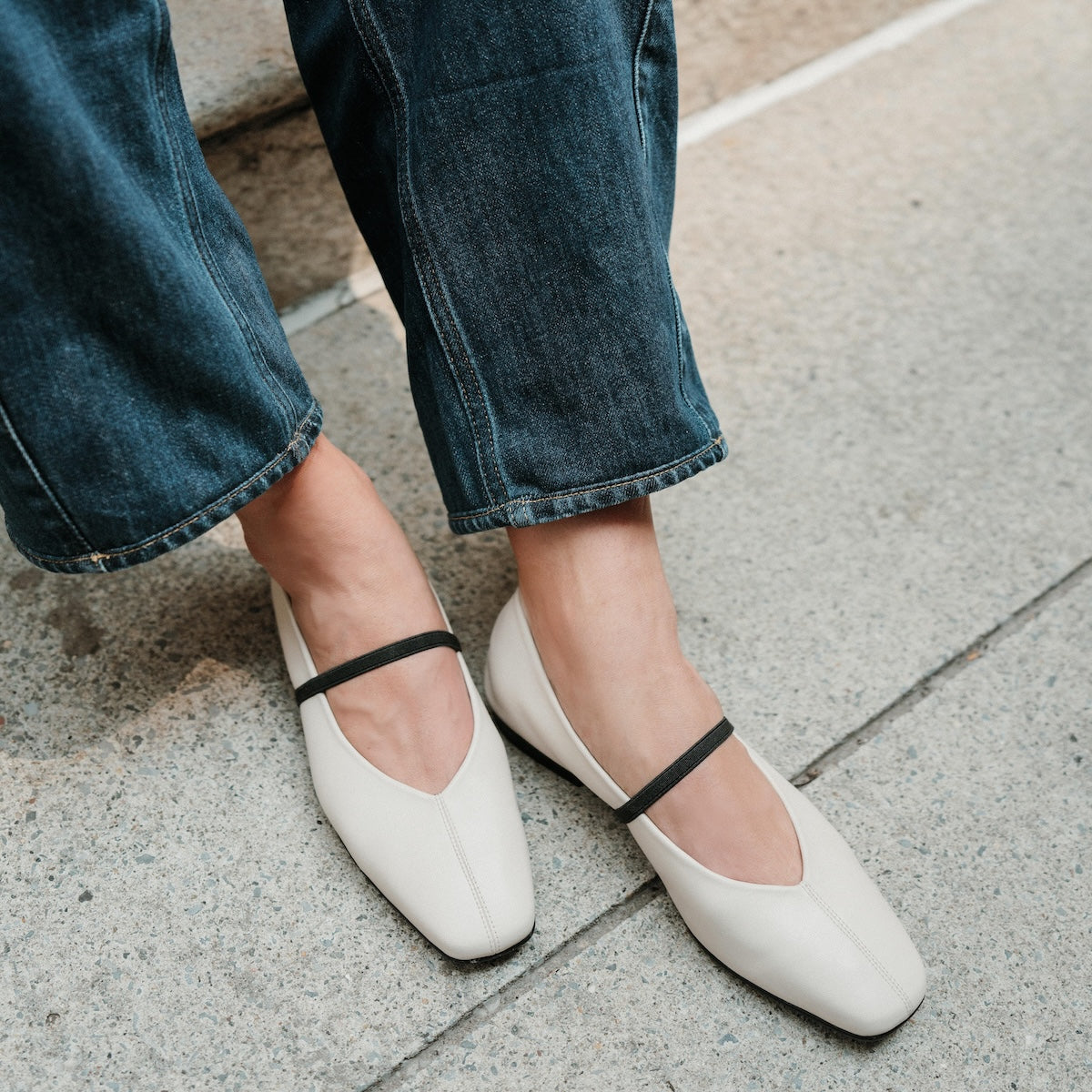 White flats with black straps worn with blue jeans on a concrete surface