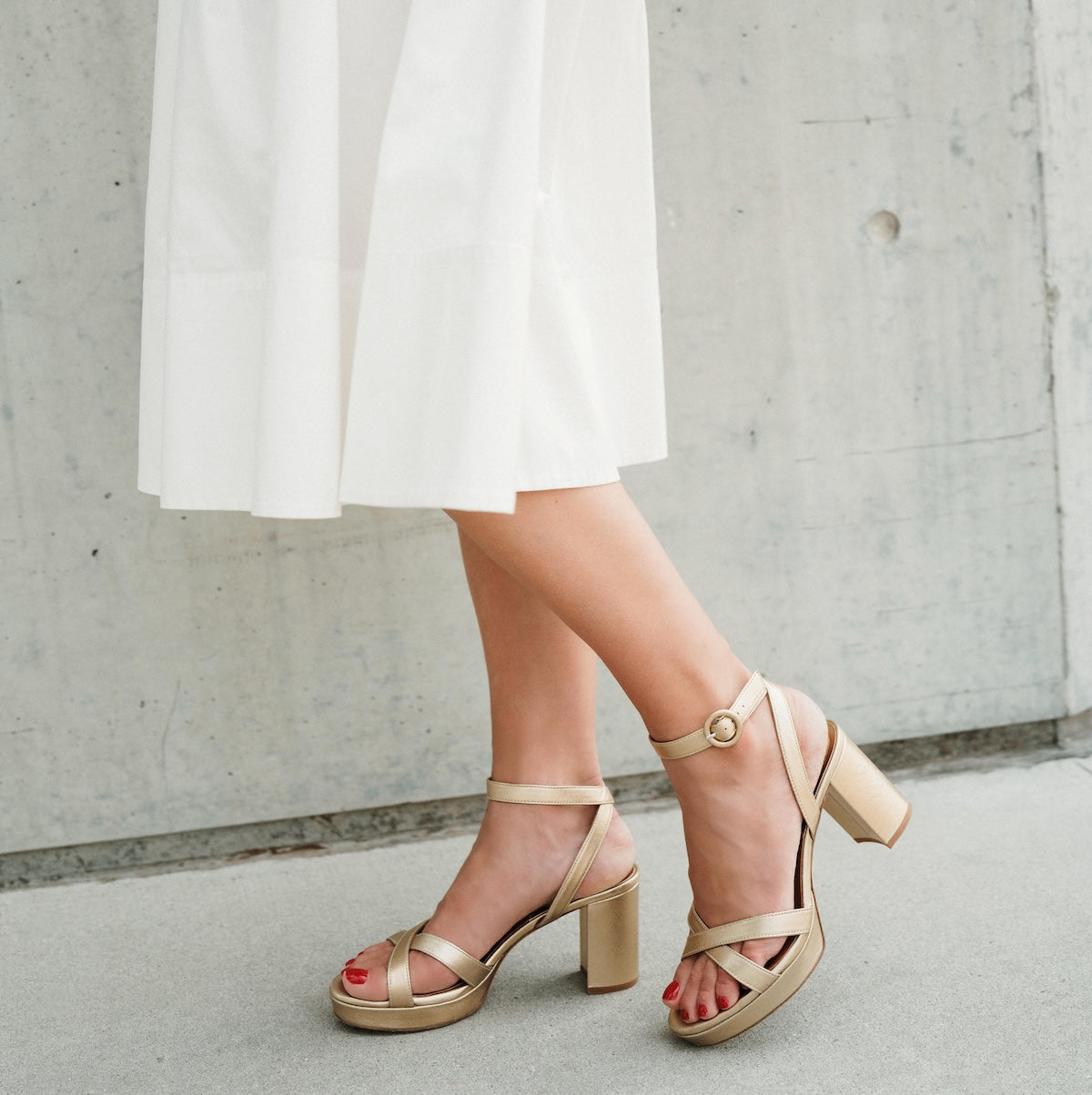 Beige sandals worn with a white skirt against a concrete wall.