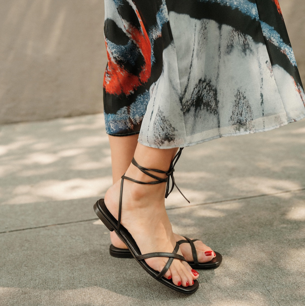 Black strappy sandals worn with a colorful skirt on a pavement background