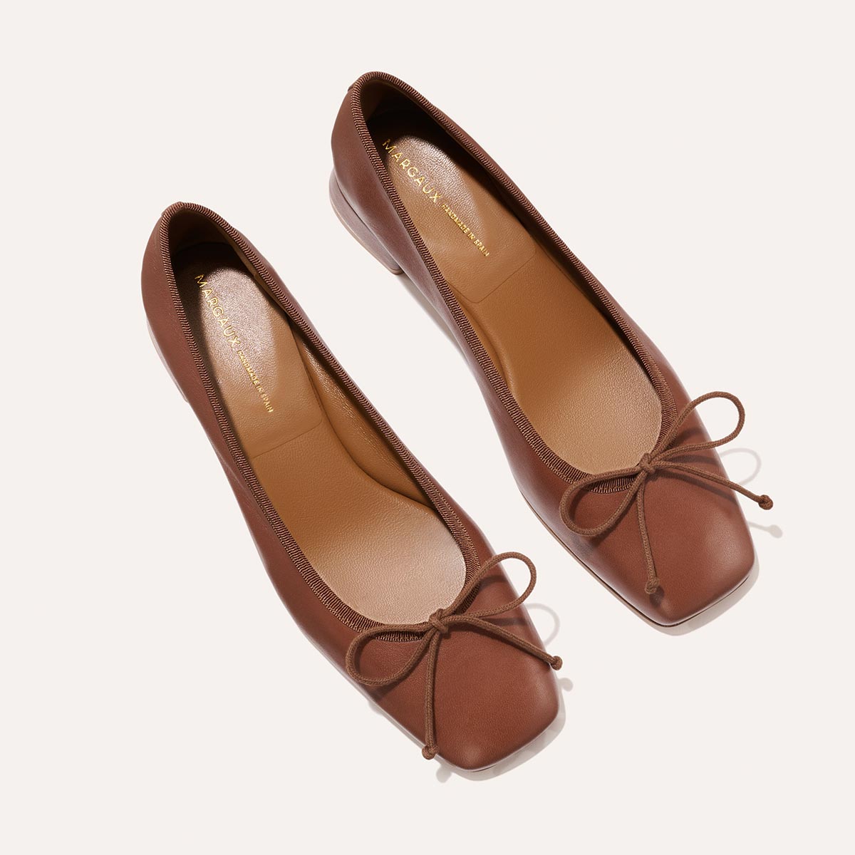 The Ada - Cognac Nappa by margaux-development: brown leather ballet flats with square toes and delicate bows, made using sacchetto construction for a glove-like fit, shown on a light background.