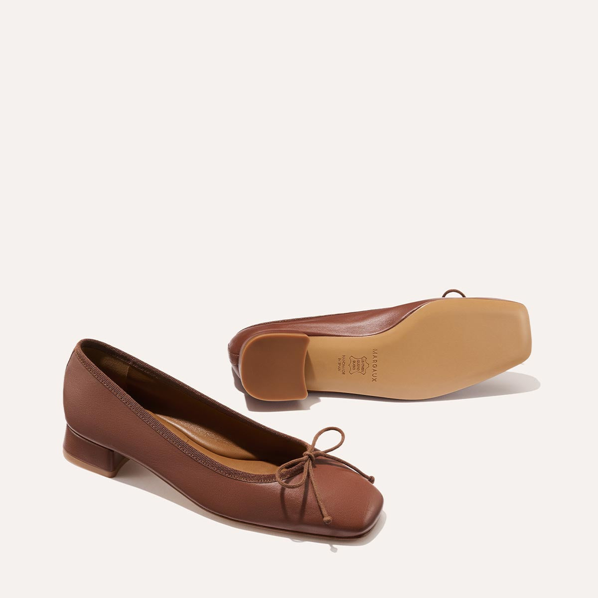 The Ada - Cognac Nappa by margaux-development: brown leather ballet pumps with low heels, square toes, bow detail, and a glove-like fit—one shoe stands upright while the other lies on its side to show the sole.