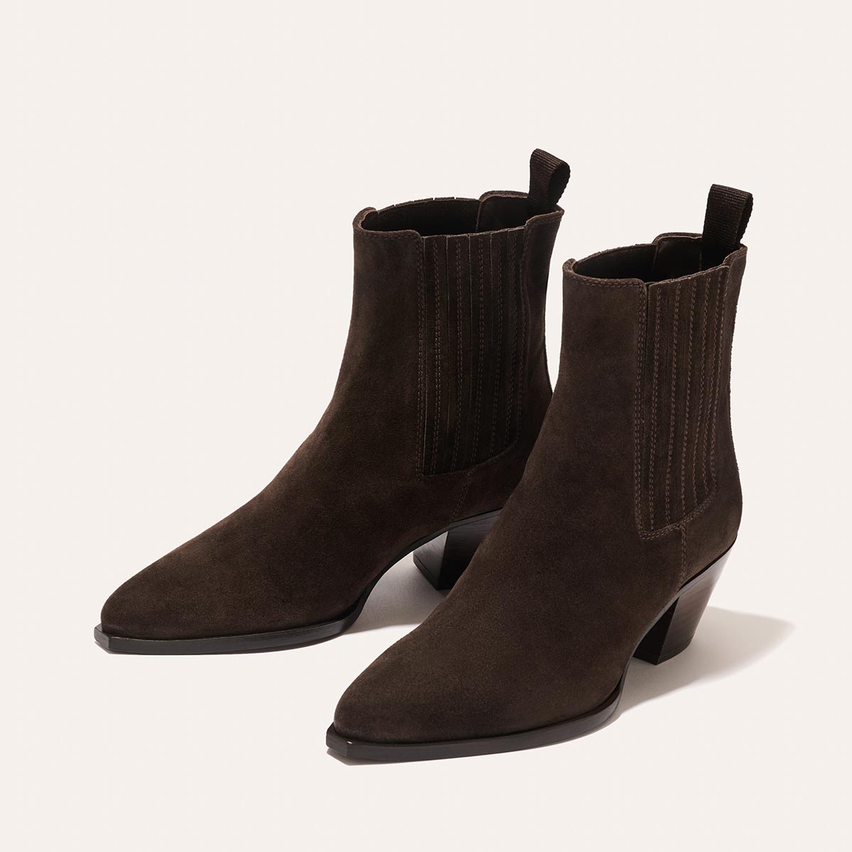 The Agnes Boot - Cocoa Suede by margaux-development: dark brown suede Western ankle boots with pointed toes, elastic side panels, pull tabs, squared-off welt, and mid-height angular heels, shown on a light background.