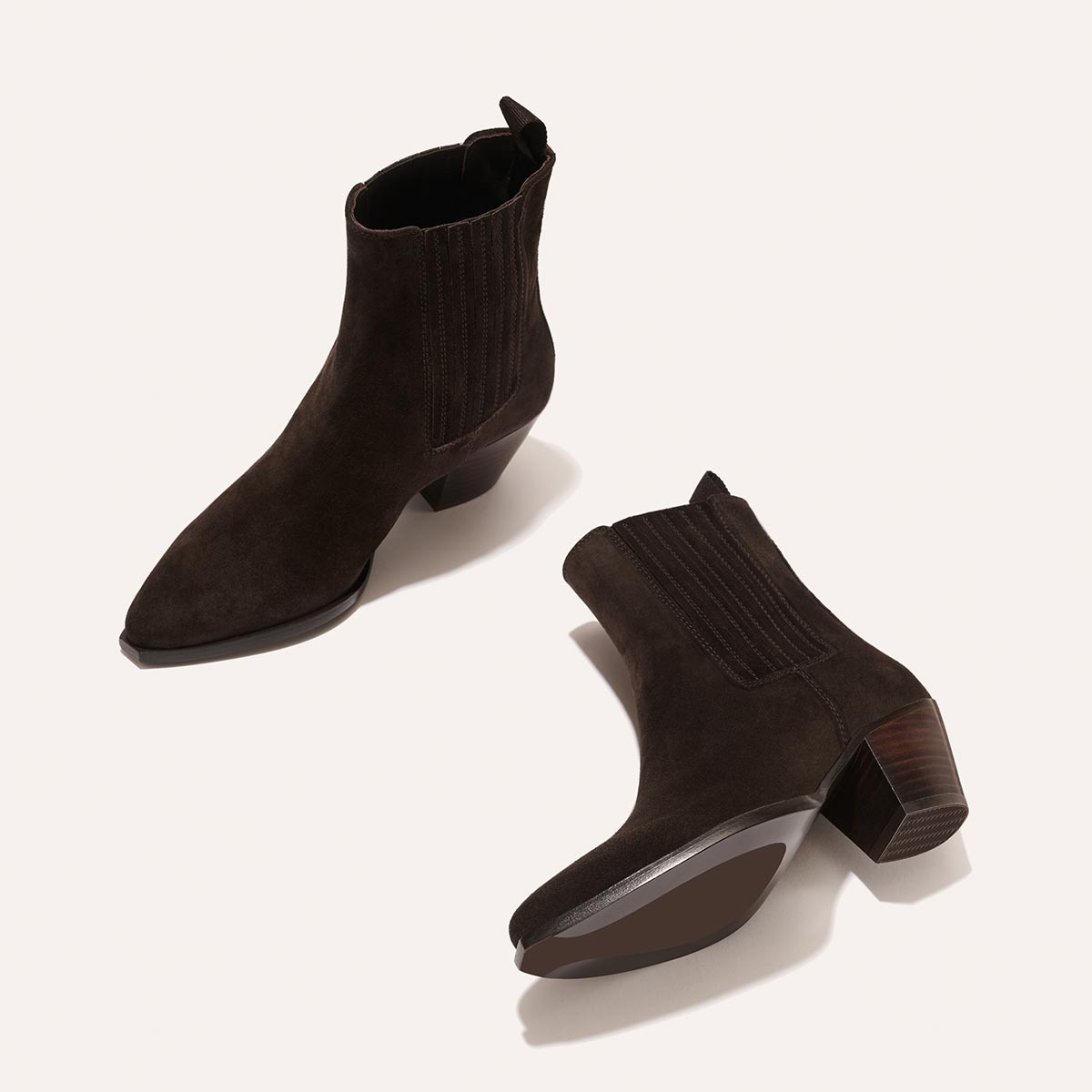 The Agnes Boot - Cocoa Suede by margaux-development: dark brown suede Western ankle boots with pointed toes, chunky angular heels, elastic side panels, and a squared-off welt. One boot stands upright; the other lies on its side on a light background.