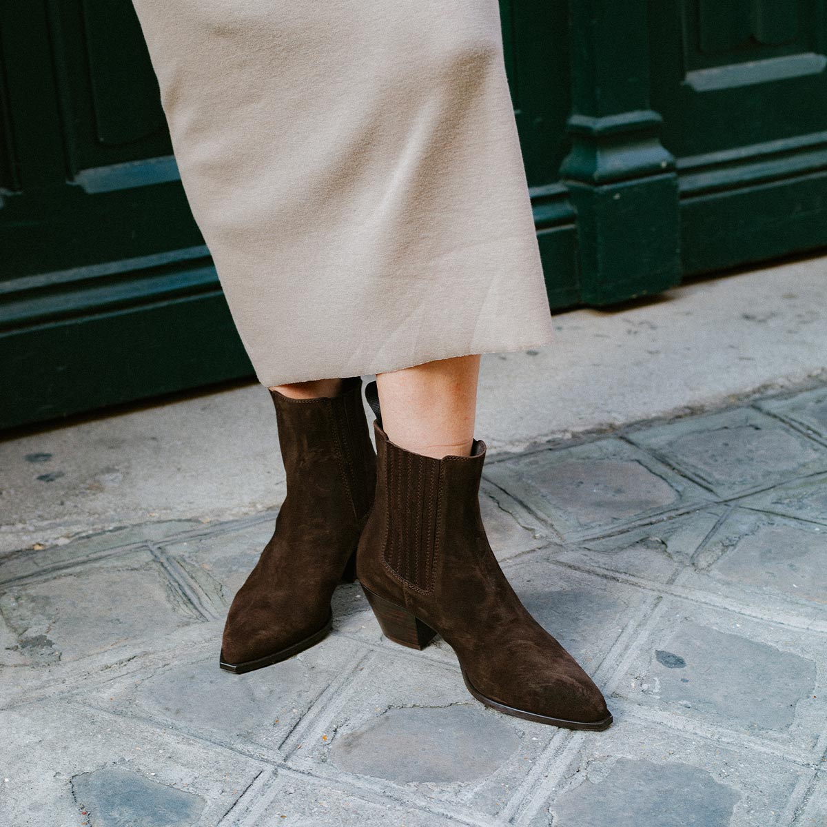 A person wearing a beige skirt and The Agnes Boot - Cocoa Suede by margaux-development stands on a stone pavement in front of a green door.