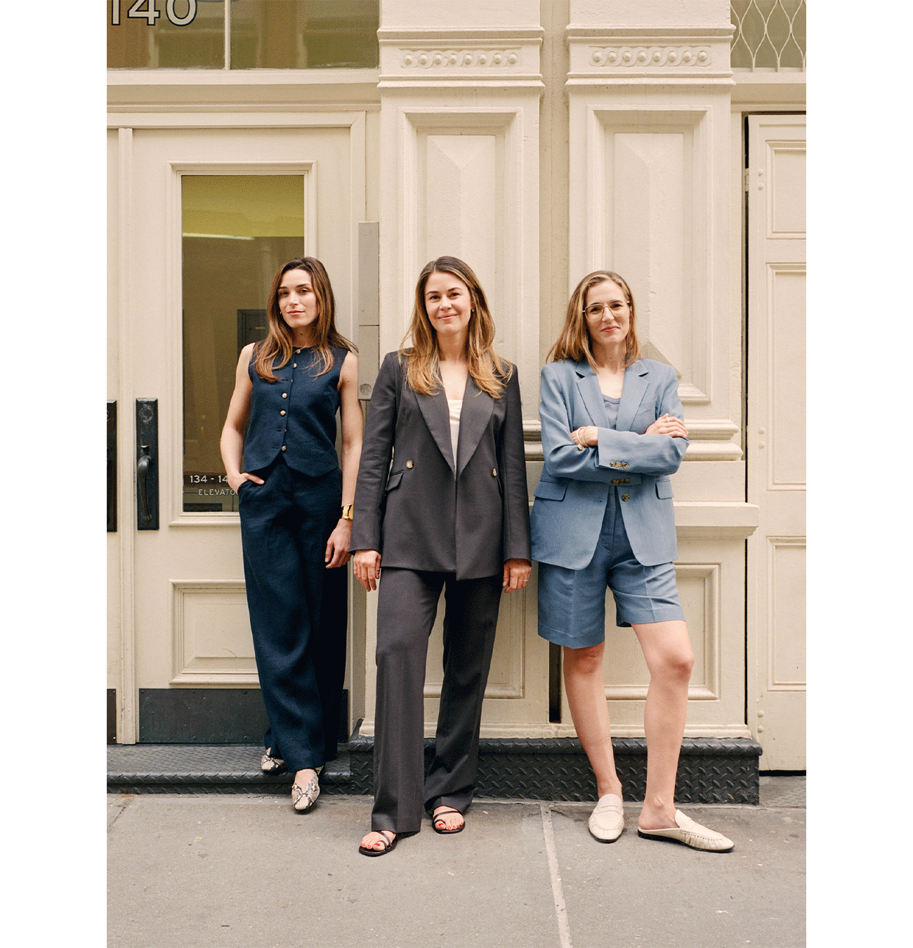 Three women stand in front of a tan building entrance, each wearing stylish business attire: a dark vest and pants, a dark double-breasted suit, and a blue blazer with matching shorts and loafers. They look confidently at the camera.