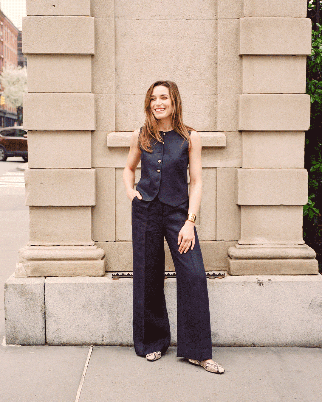 A woman wearing a sleeveless navy vest and matching wide-leg pants stands confidently in front of a stone building, smiling with one hand in her pocket. She has long brown hair and wears snakeskin shoes.