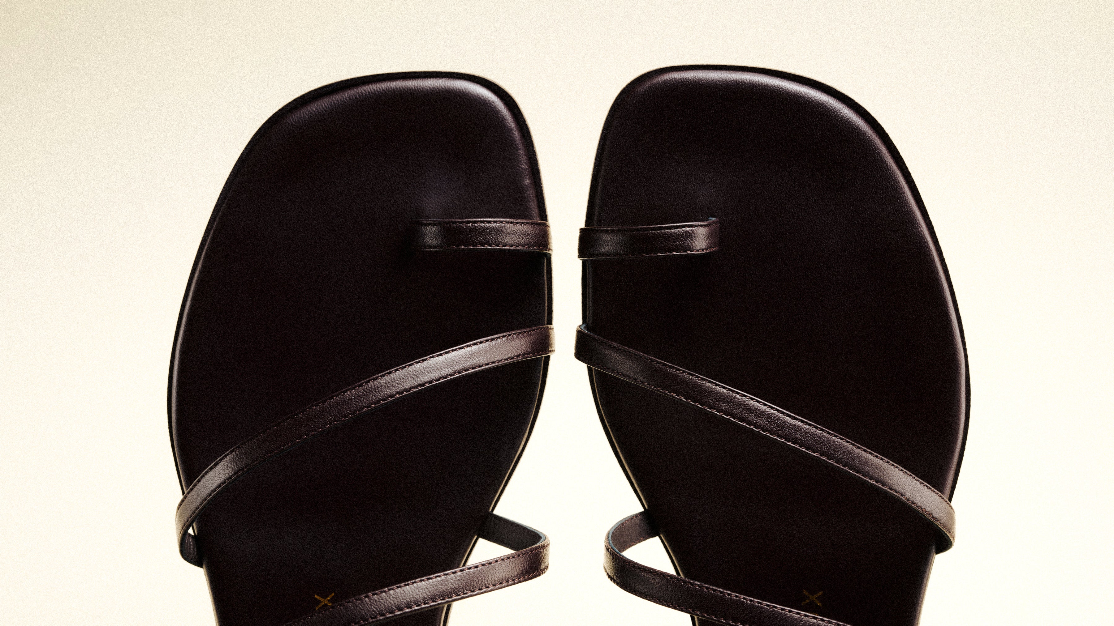 A close-up view of a pair of dark brown flat sandals with square toes and thin crossing straps, set against a light background.