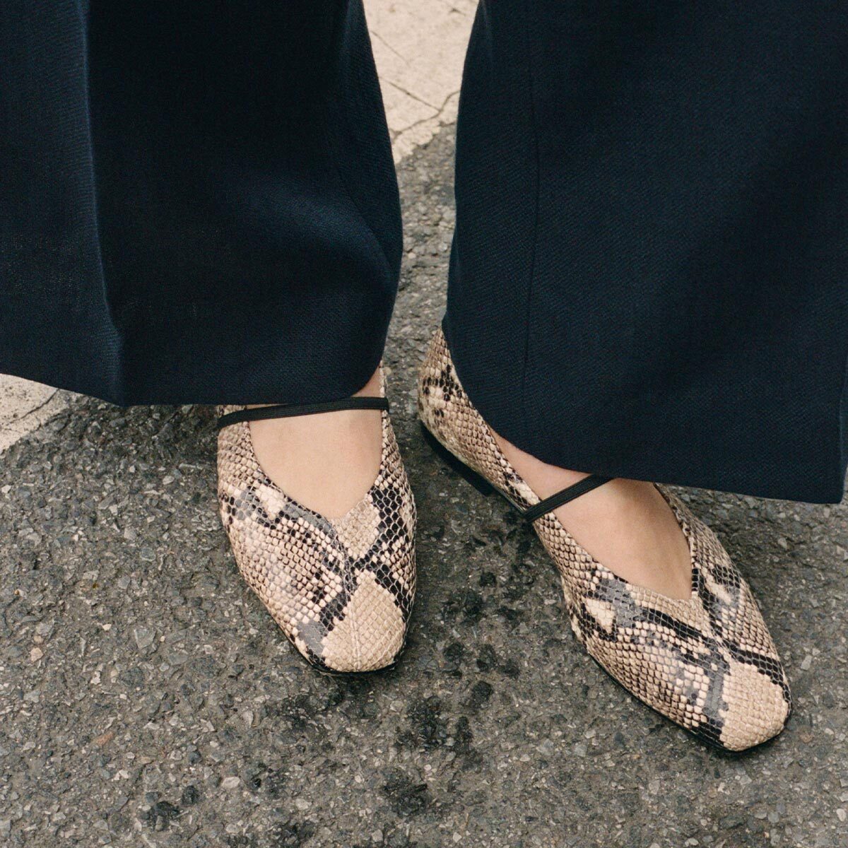 A person wearing black pants and The Phoebe - Natural Python Embossed flats by margaux-development with thin straps stands on textured pavement.