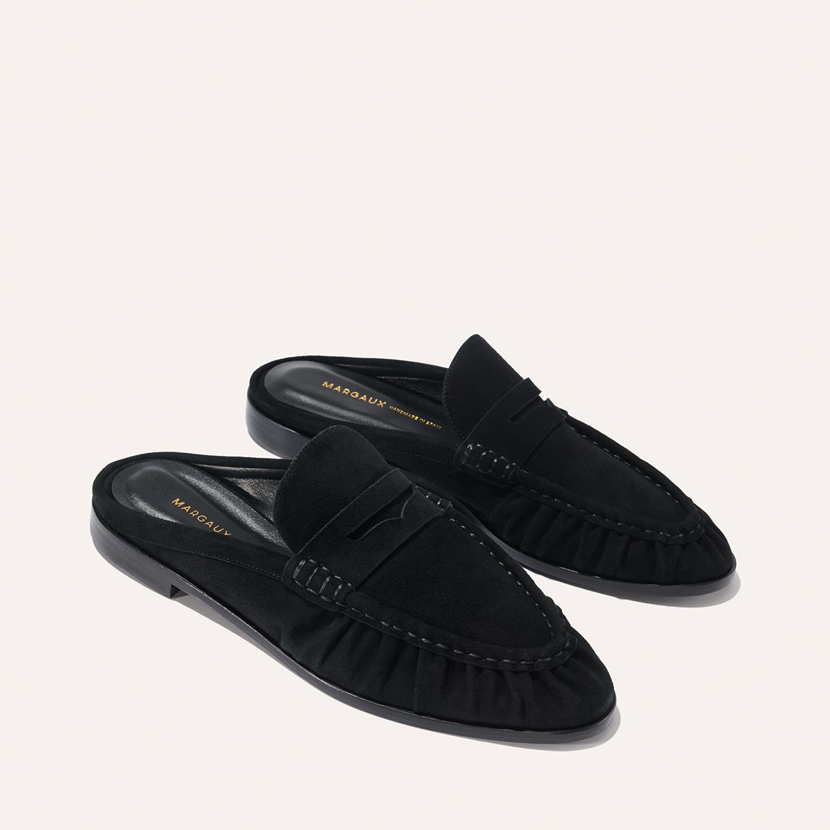 The Margaux Astor Loafer Mule in black suede features a low heel and ruched side detailing, showcased against a light background.