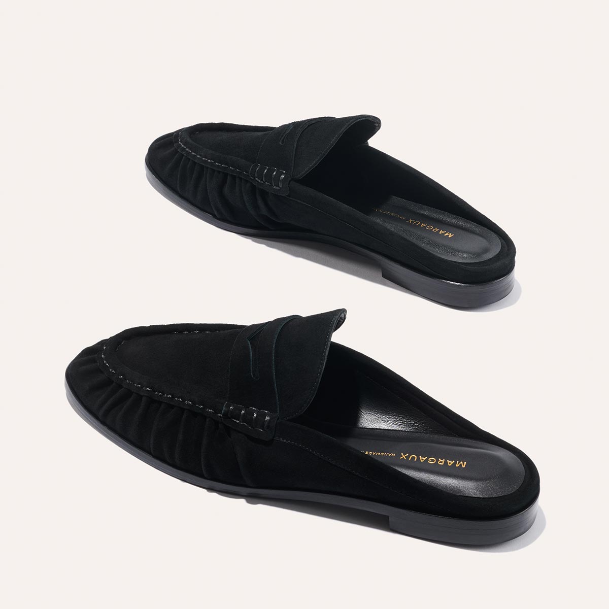 A pair of The Astor Loafer Mule in black suede by Margaux, featuring ruched sides and flat soles, is displayed on a light background. The insoles are branded with Margaux.