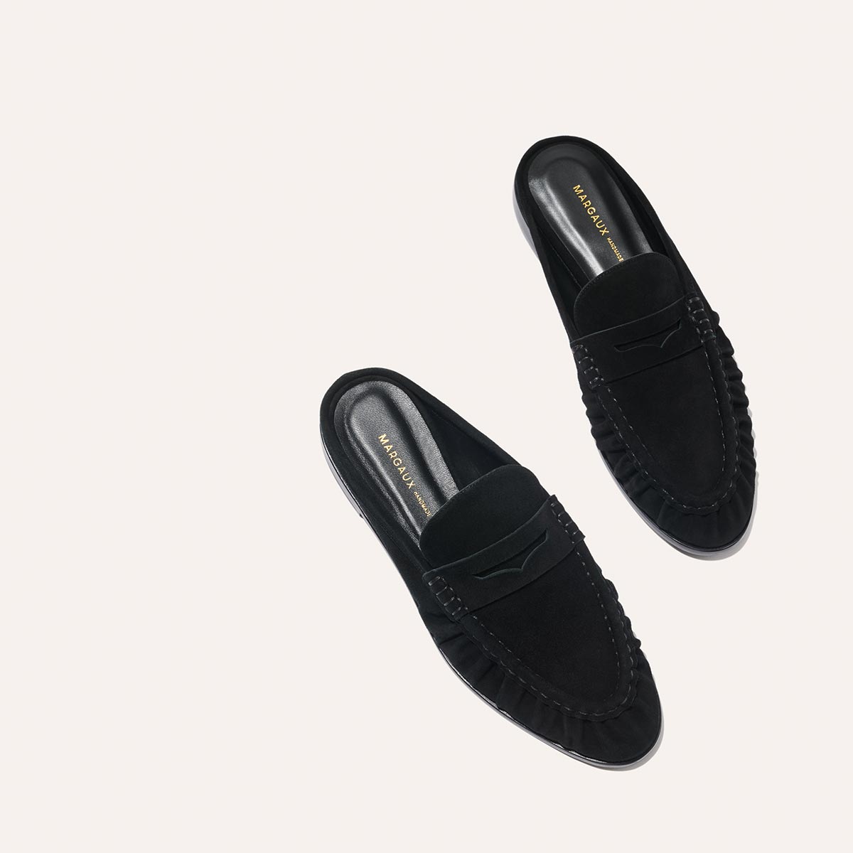 The Astor Loafer Mule by Margaux in black suede features stitching details and gold branding on the insoles, shown against a light beige background.