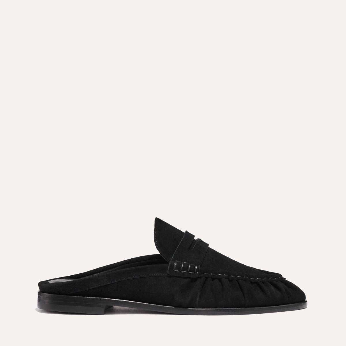 The Astor Loafer Mule by Margaux in black suede features a backless design, subtle ruching near the sole, visible stitching, and a low flat heel, shown in side view against a plain light background.