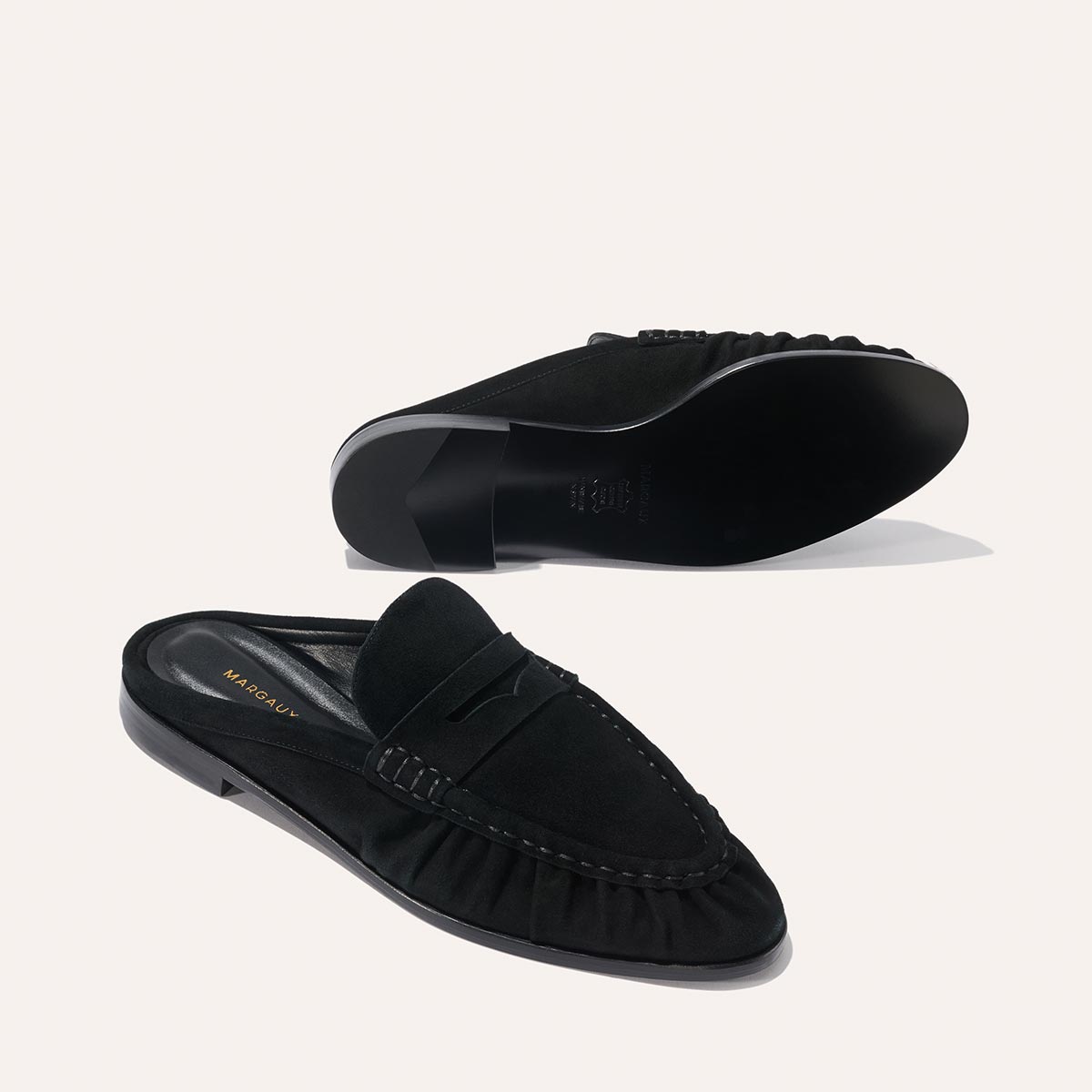 The Astor Loafer Mule in black suede by Margaux features a low heel and ruched sides; one shoe stands upright while the other shows the sole, both displayed on a light background.