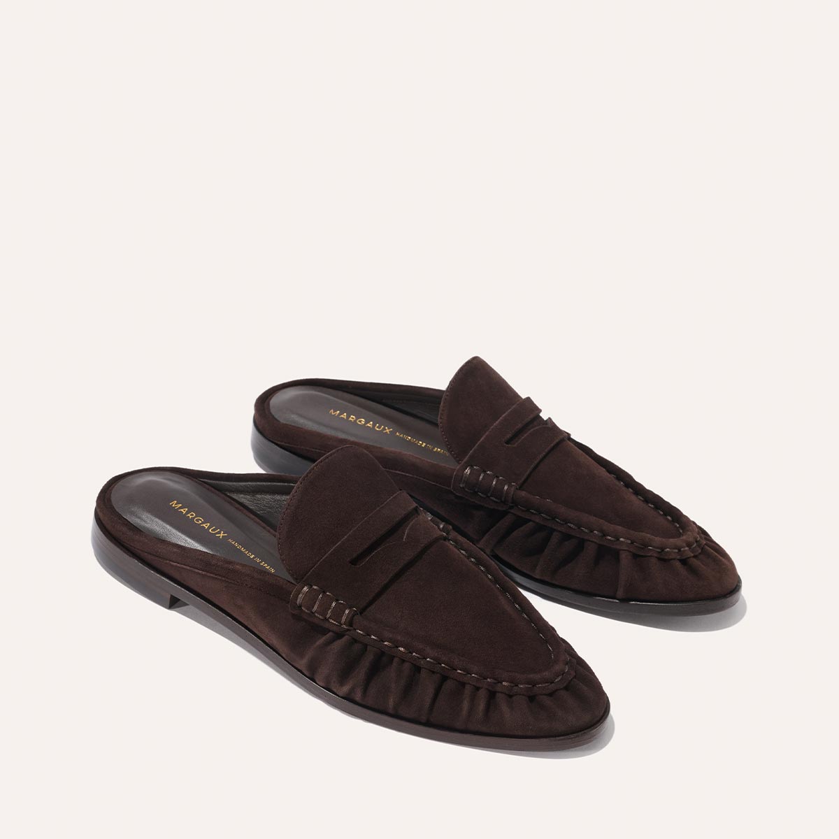 The Astor Loafer Mule in Espresso Suede by margaux-development features low heels, decorative stitching, and pleated side detailing. Displayed against a plain light background.