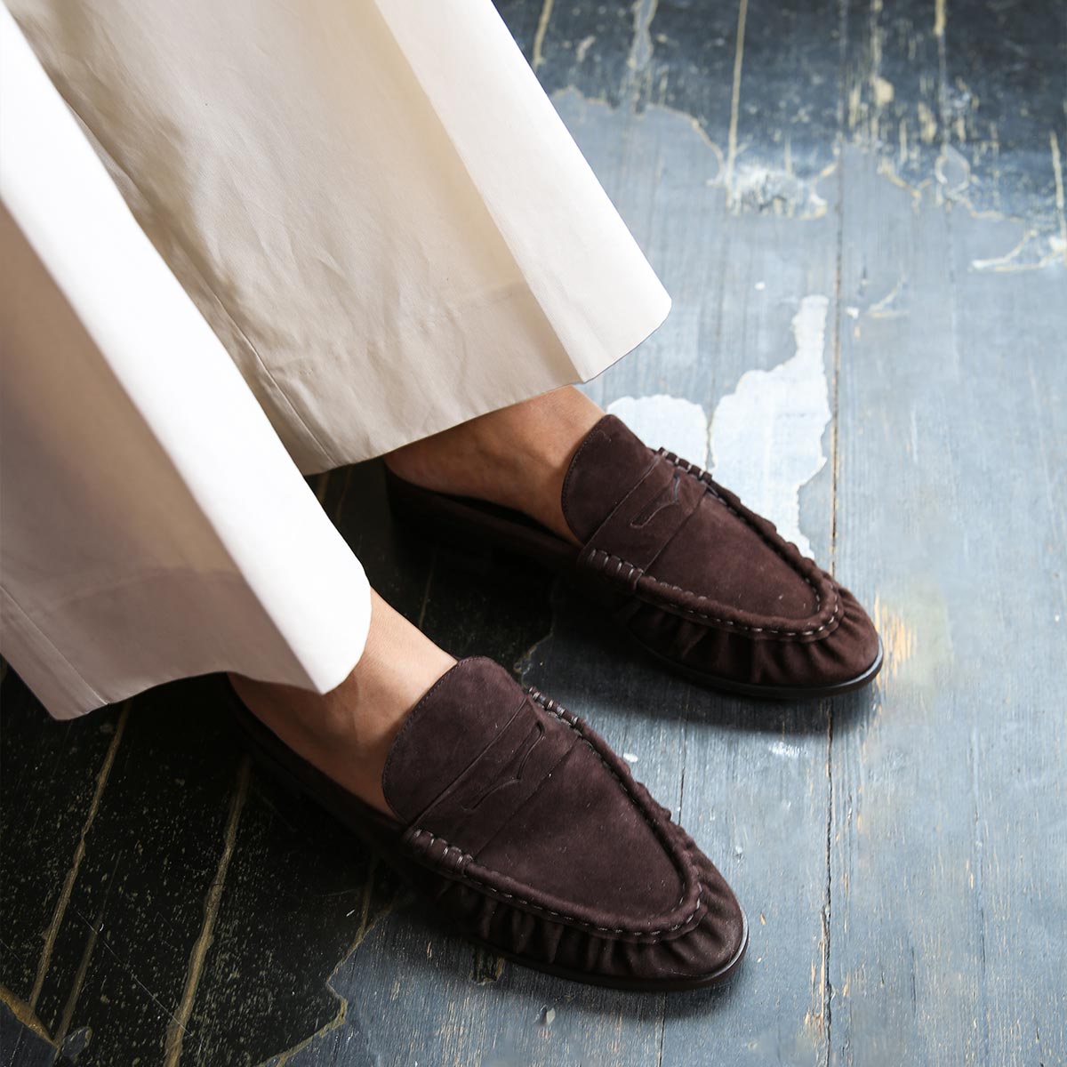A person wearing cream-colored wide-leg pants and margaux-developments The Astor - Espresso Suede stands on a worn, painted wooden floor.
