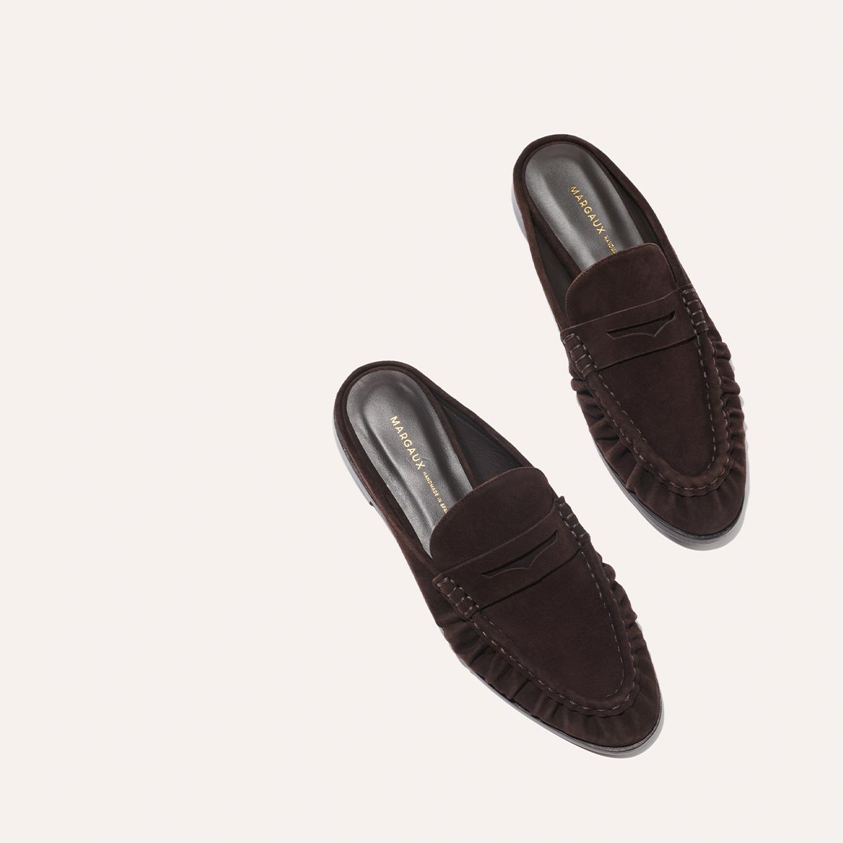 The Astor Loafer Mule in Espresso Suede by margaux-development features a slip-on design, visible stitching, and a penny slot detail. Shown from above on a light background.