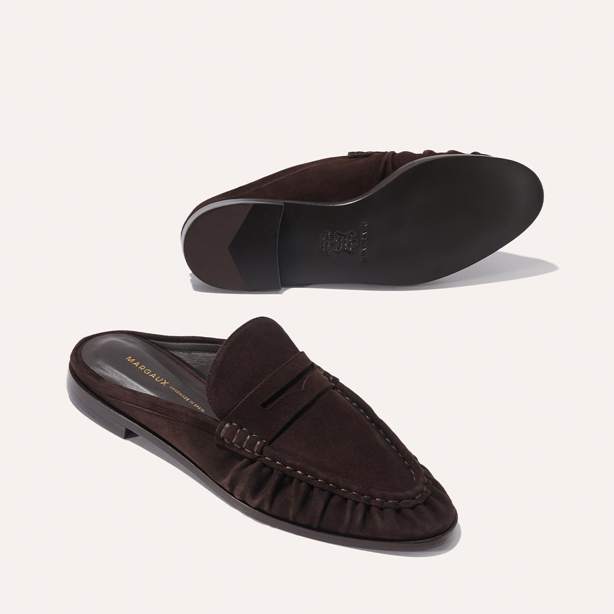 The Astor Loafer Mule in Espresso Suede by margaux-development, featuring ruched detailing, a flat leather sole, and subtle stitching. One mule is shown from the top and the other from the sole on a light background.