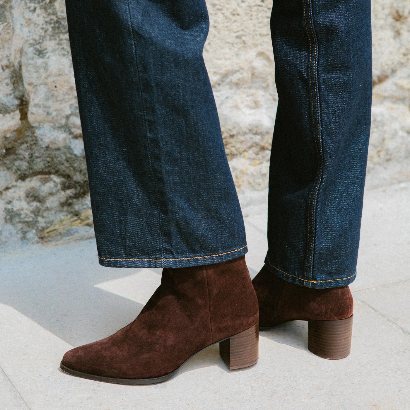 A person wearing dark blue jeans and brown suede ankle boots with a block heel stands on a light-colored sidewalk near a stone wall.