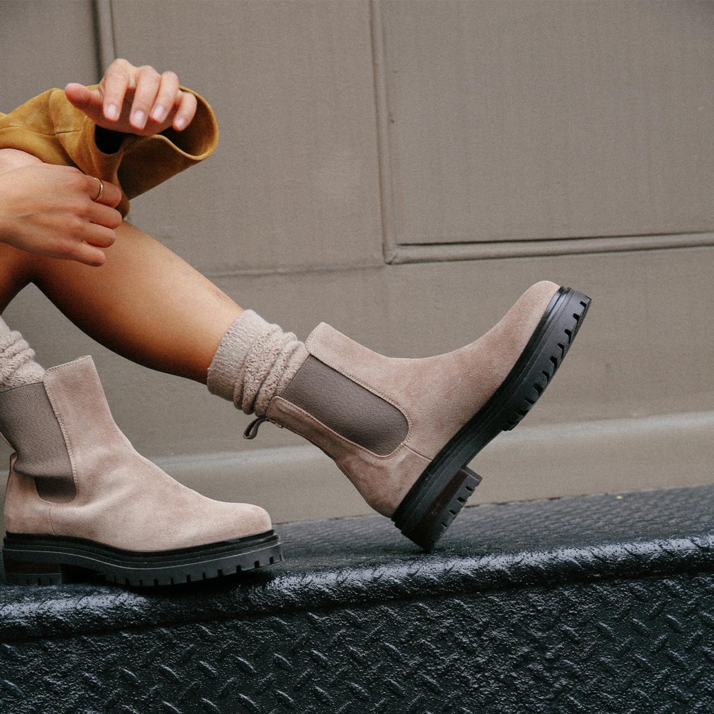 A person wearing beige suede Chelsea boots and scrunched socks sits on a textured black metal surface, with their legs extended and an arm resting on their knee against a neutral wall background.