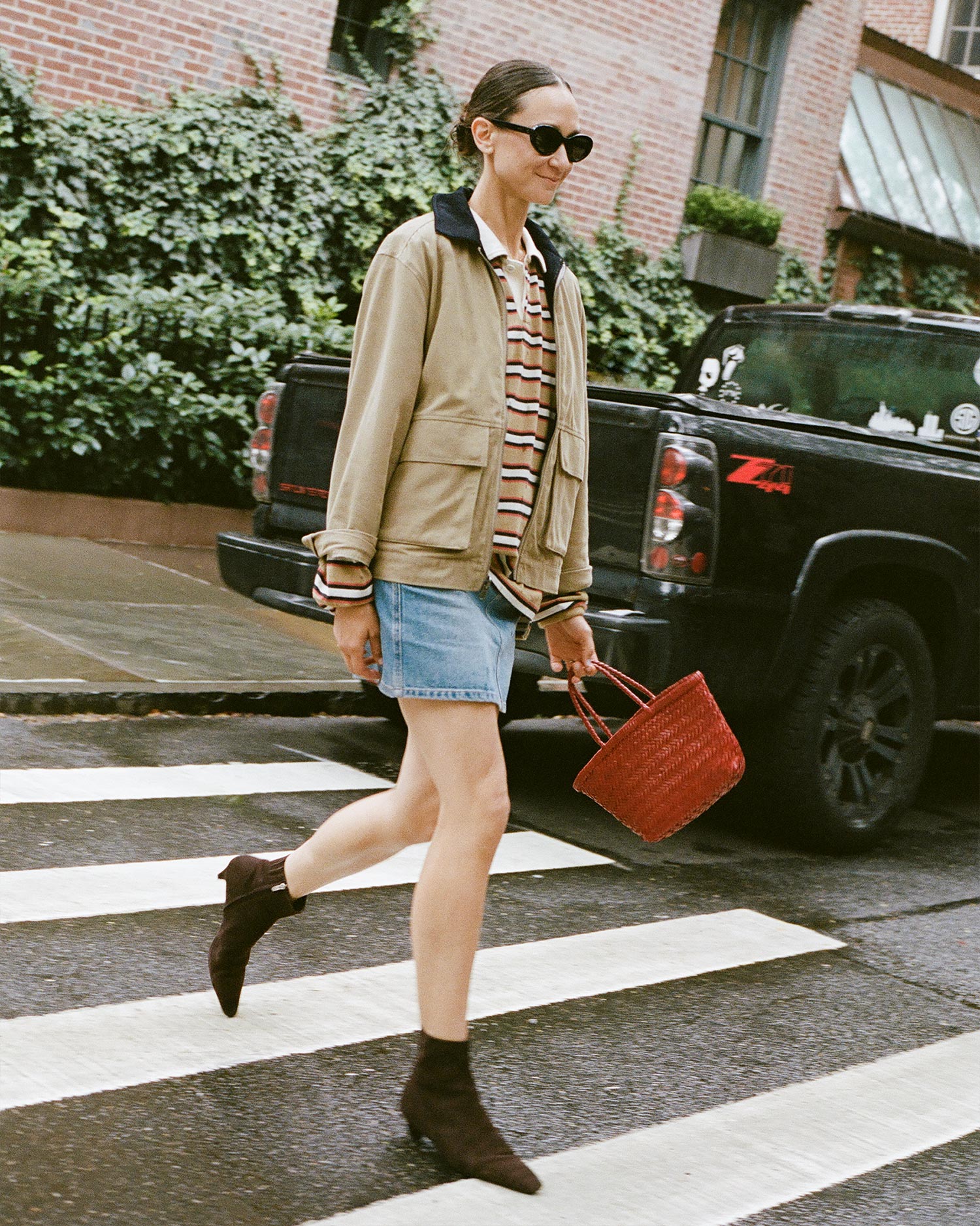 A woman wearing sunglasses, a beige jacket, a striped shirt, a denim skirt, and ankle boots walks across a crosswalk holding a red woven bag, with a black pickup truck and brick building in the background.