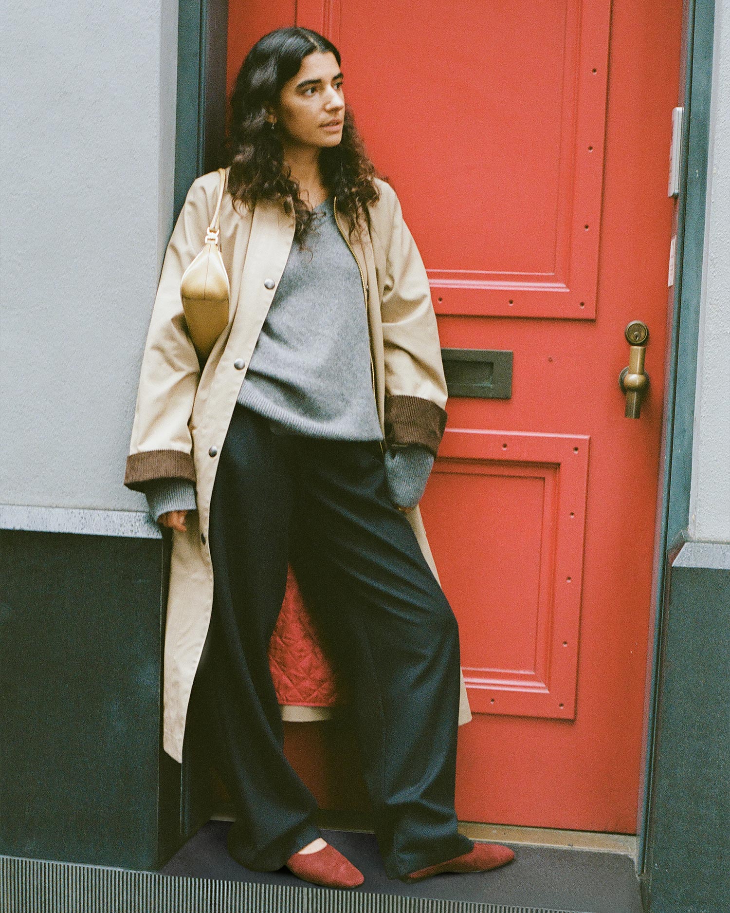 A person with long dark hair stands in front of a red door, wearing a beige trench coat, grey sweater, black wide-leg pants, and red shoes, with a tan bag over one shoulder.