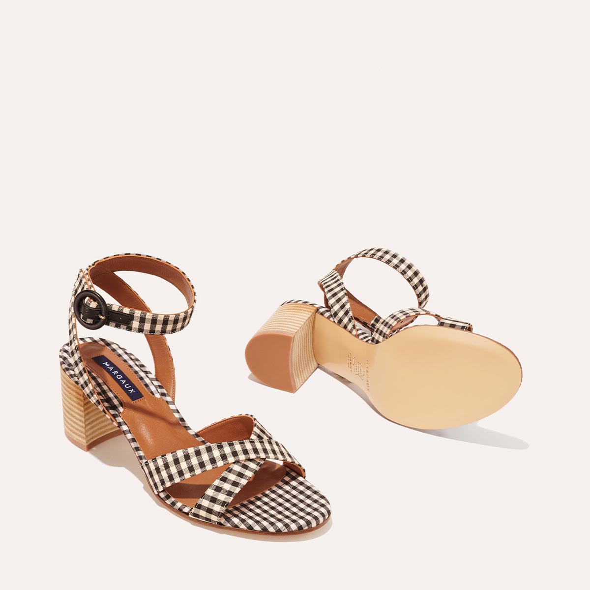 The City Sandal by margaux-development features tan block heels, a black and white gingham pattern, ankle buckled straps, and cushioned insoles for comfort. One sandal stands upright while the other displays its sole.