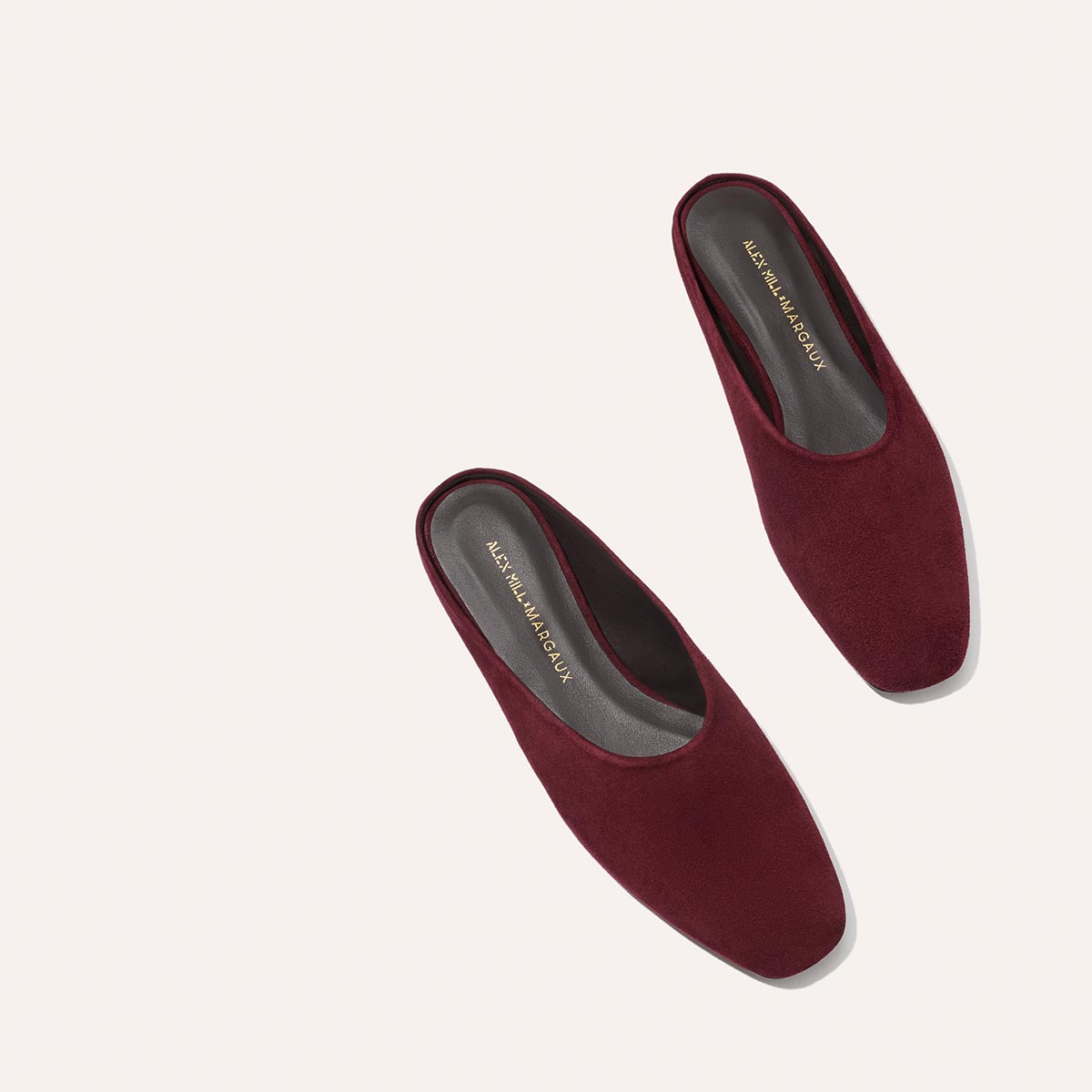 The Alex Mill x Margaux: The Clara - Burgundy Suede by Margaux is a pair of square-toe suede mule flats in burgundy, shown on a light beige background with dark insoles featuring gold text.