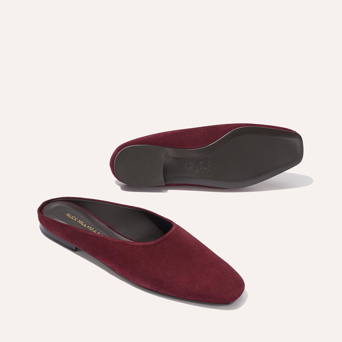 The Margaux Alex Mill x Margaux: The Clara - Burgundy Suede flats feature a closed, rounded toe and black sole; one stands upright while the other lies on its side, showcasing their stylish versatility.