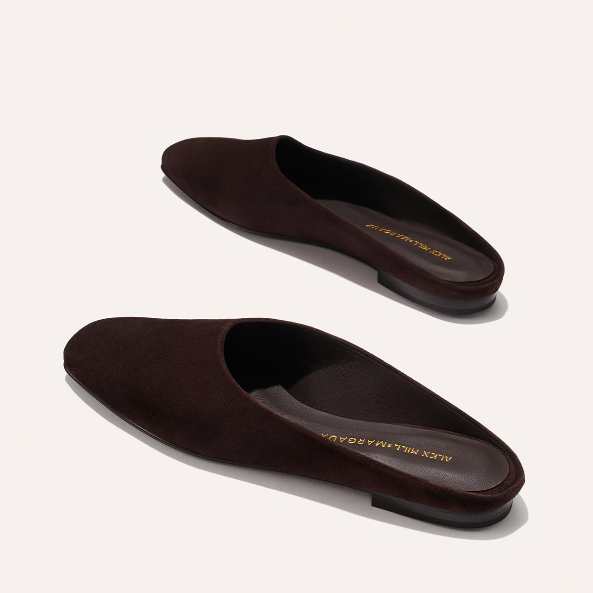 The Clara - Espresso Suede by Margaux: dark brown suede slip-on mules with a slipper-inspired silhouette and flat soles, featuring gold Alex Mill x Margaux branding on the insoles and shown on a light beige background.