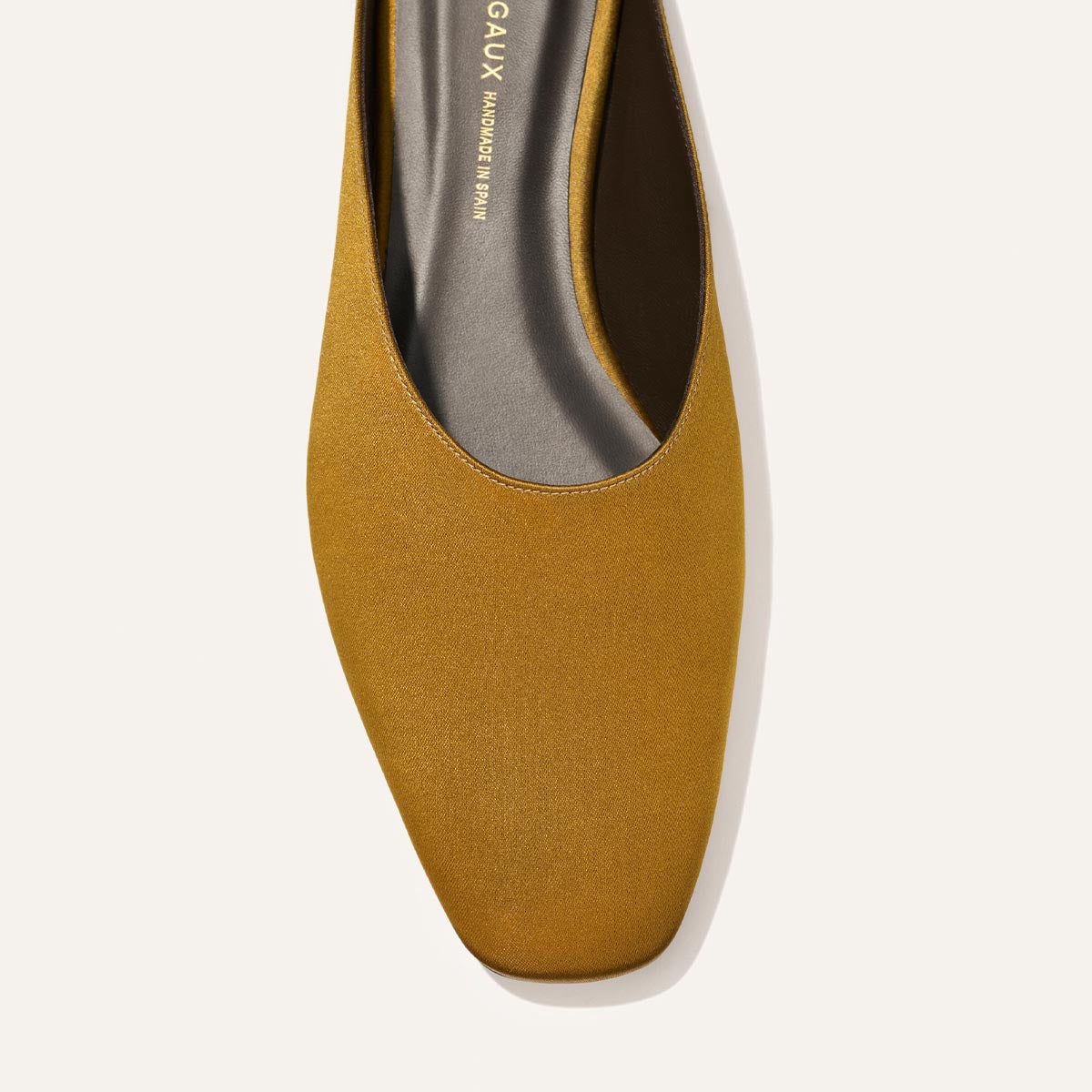 A close-up top view of The Clara - Ochre Satin flat by Margaux, featuring a square toe and smooth satin finish, set against a light beige background.
