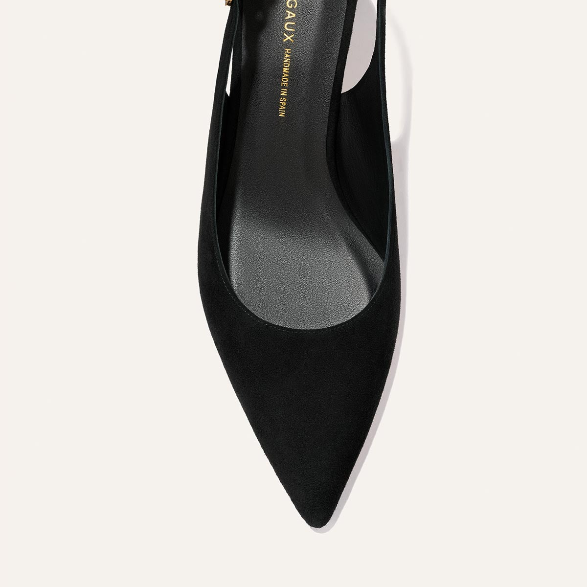 A close-up top view of Margaux's The Colette - Black Suede high heel showcases its pointed toe, smooth suede finish, and gold branding on the insole against a light, neutral background.