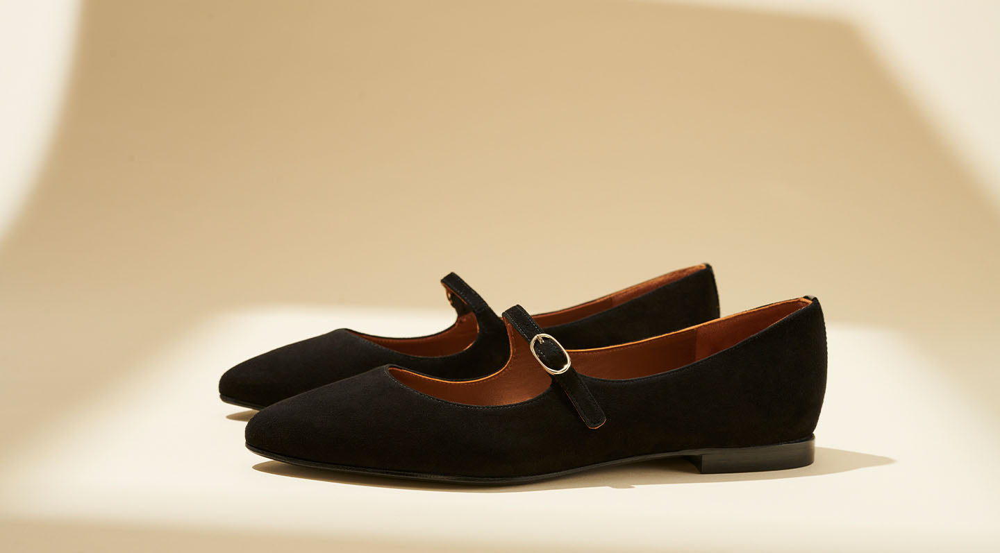 A pair of black suede Mary Jane flats with a thin strap and small buckle, displayed on a neutral background with soft lighting and shadows.