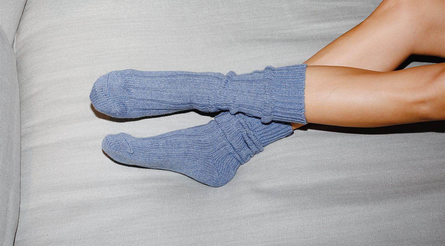A person with bare legs is lying on a light gray couch, wearing thick, light blue knitted socks.