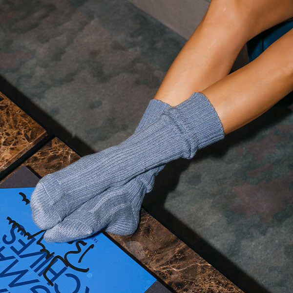 A person with crossed legs wearing light blue ribbed socks rests their feet on a dark brown marble table, with a blue and black magazine visible in the foreground.