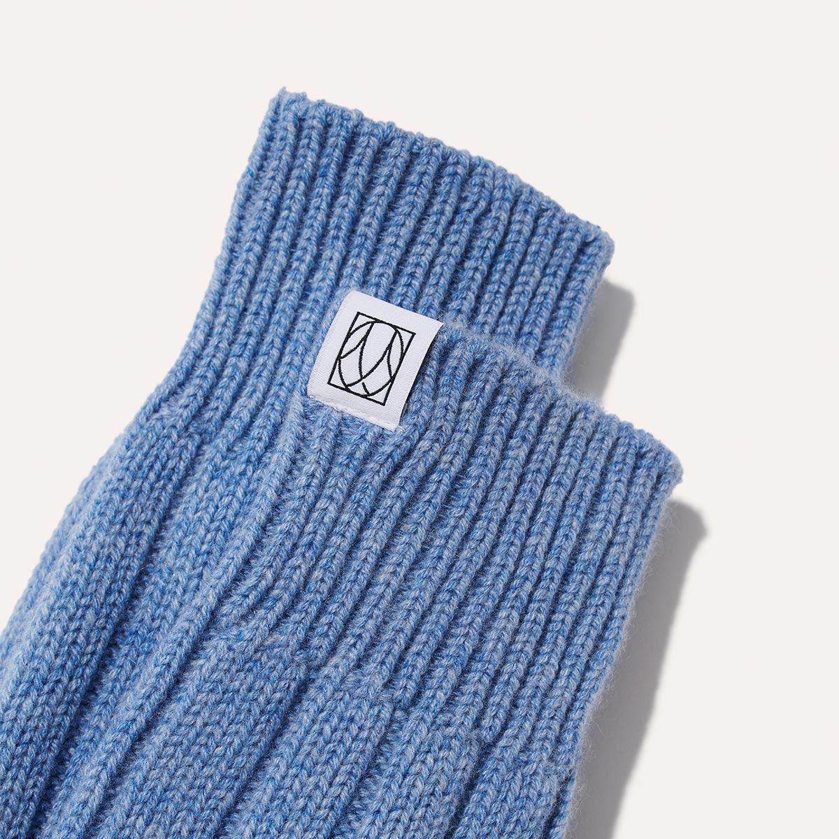 A close-up of Margauxs The Cozy Sock in light blue wool-cashmere blend, featuring a ribbed knit and a white patch with black logo near the cuff—luxurious comfort for lounging, shown on a light background.