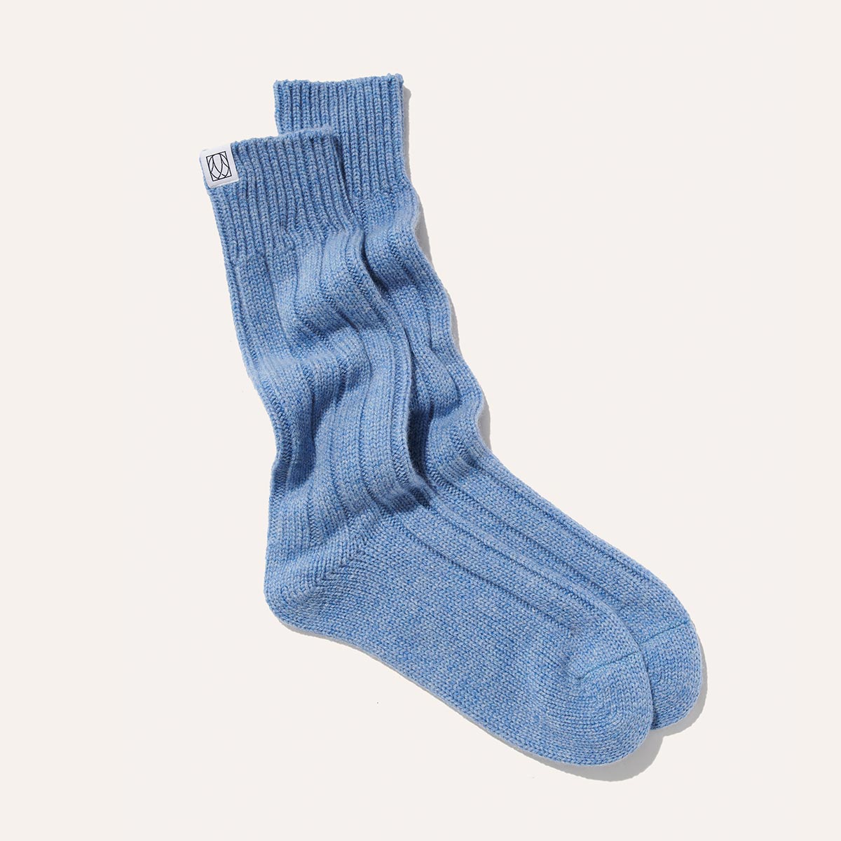 The Cozy Sock by Margaux in light blue wool/cashmere blend is laid flat on a white background, featuring ribbed texture and a white tag with black logo near the cuff—ideal for luxurious lounging.
