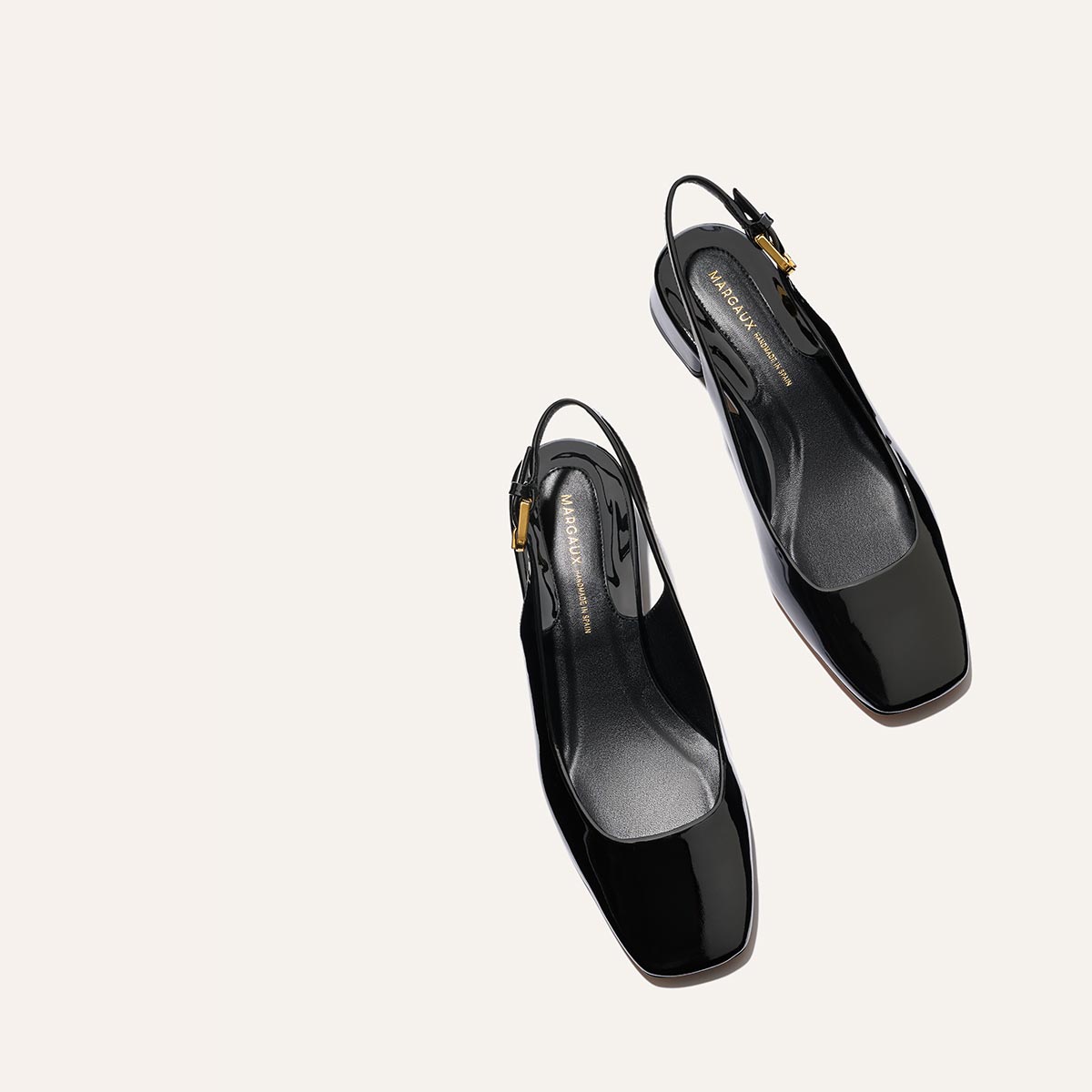 The Darcy Slingback - Black Patent by Margaux features adjustable slingback straps, square toes, low heels, and gold-tone buckles. These elegant patent leather ballet pumps are shown on a light beige background for a refined look.