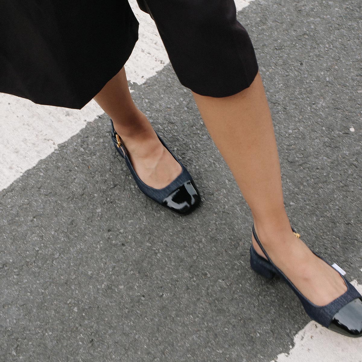 Wearing black cropped pants and Margauxs The Darcy Slingback in Indigo Denim and Patent, a person crosses a street on a crosswalk, showcasing a chic warm-weather look.