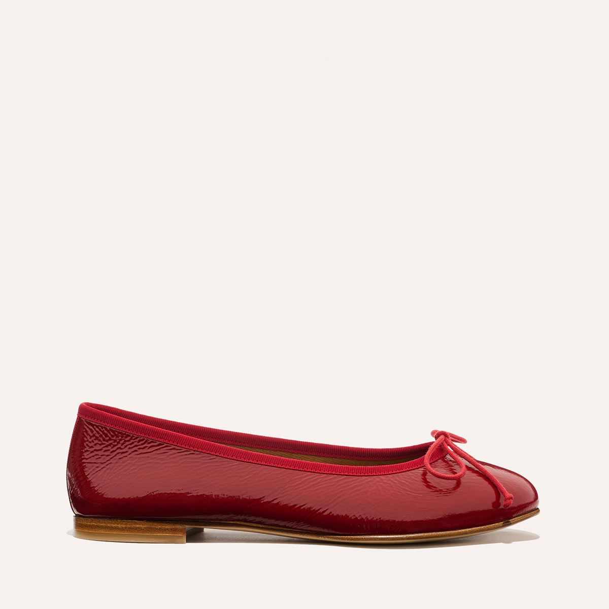 The Demi - Berry Crinkle Patent by margaux-development: a shiny red ballet flat with a small bow on the rounded toe, flat wooden sole, and glossy finish, shown on a plain light background.