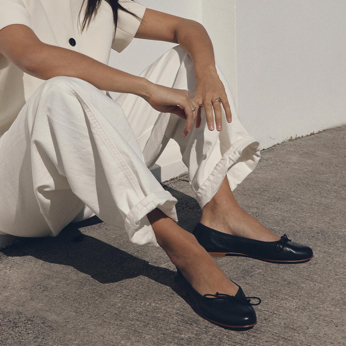 A person in white sits cross-legged on concrete, arms on knees, highlighting The Demi - Black Nappa ballet flats by margaux-development—handmade in Spain with small bows and bathed in beautiful natural sunlight.