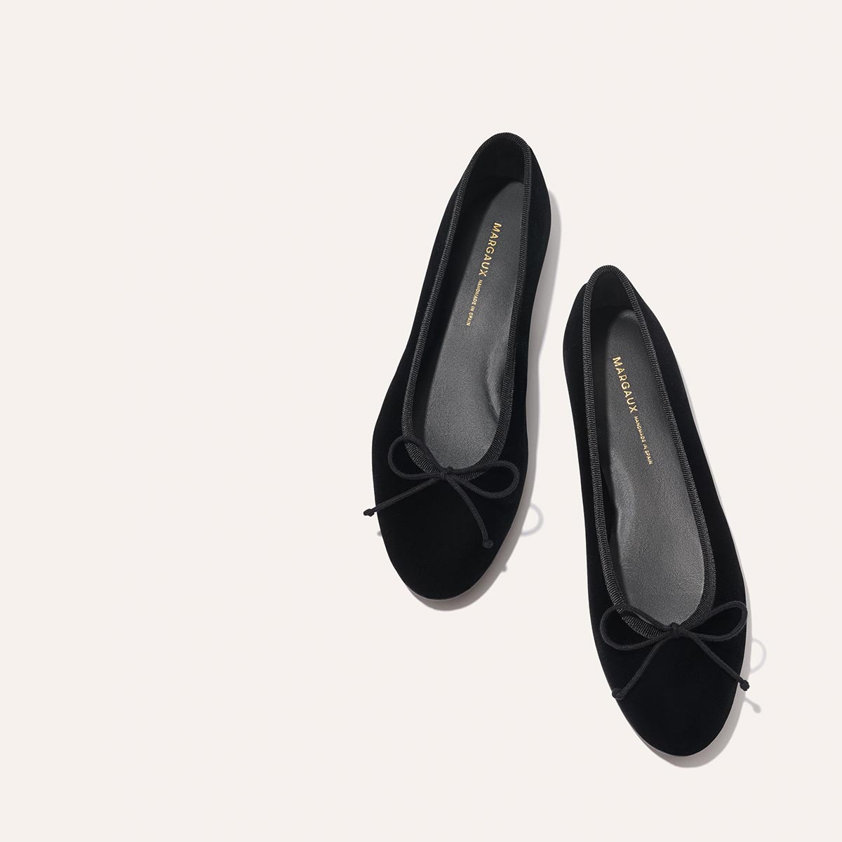 A pair of The Demi - Black Velvet ballet flats by margaux-development, featuring small bows on the toes and gray insoles with gold Margaux branding, displayed on a light background.