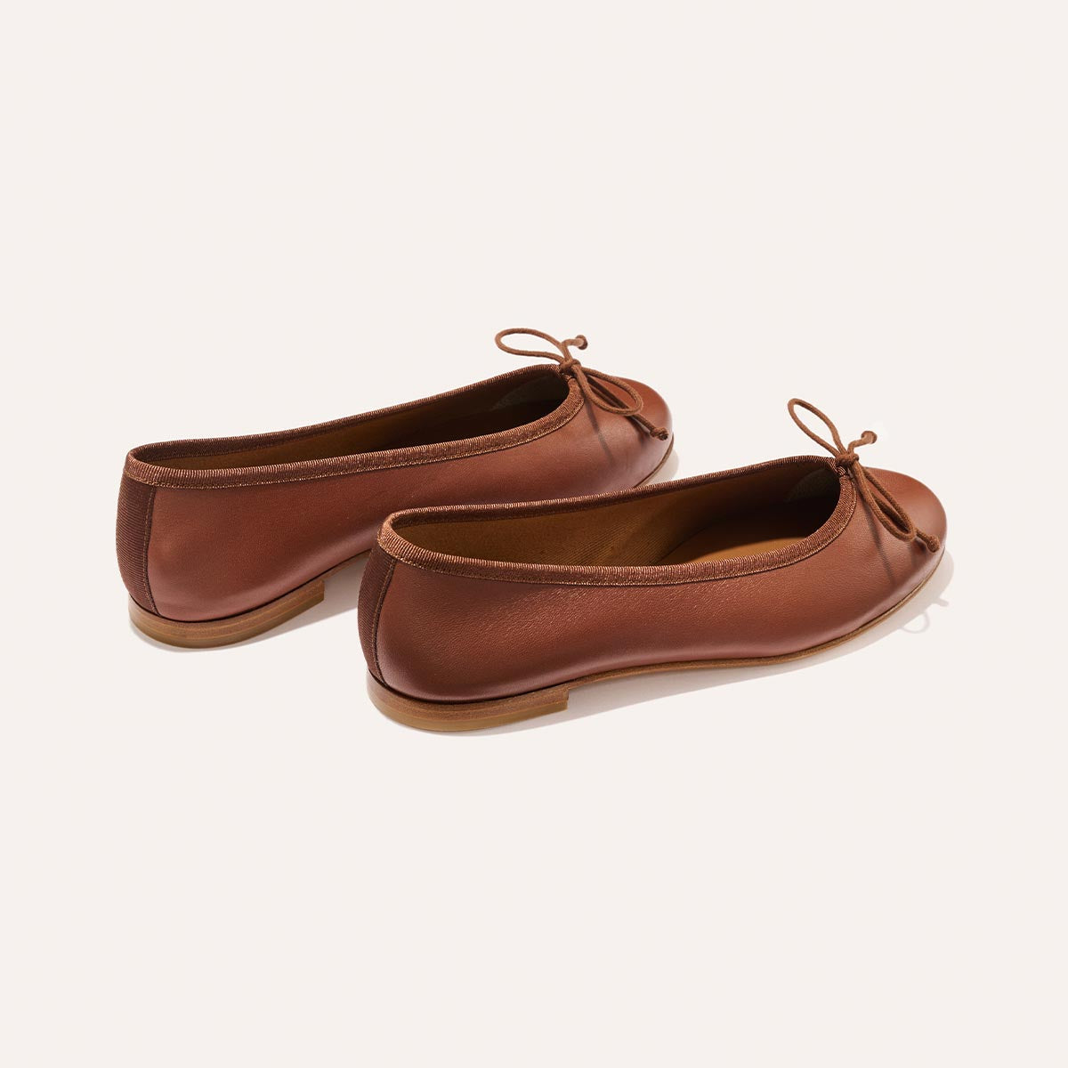 The Demi - Cognac Nappa by margaux-development: a pair of handmade brown ballet flats from Spain with small front bows and flat soles, shown against an off-white background.