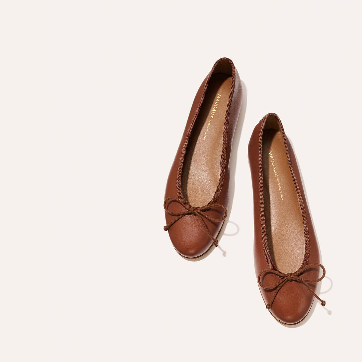 The Demi - Cognac Nappa by margaux-development: a pair of handmade brown leather ballet flats with charming toe bows, shown side by side on a light beige background.