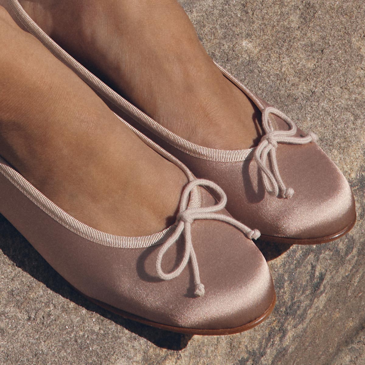 Close-up of two feet wearing The Demi - Dusty Pink Satin ballet flats by margaux-development, featuring bows, standing on a textured stone surface.