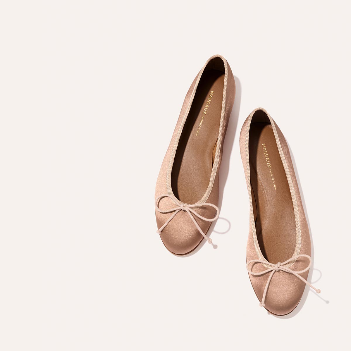 The Demi - Dusty Pink Satin by margaux-development: handmade in Spain, these light pink ballet flats feature small bows on the toes and are shown against a plain, light background.