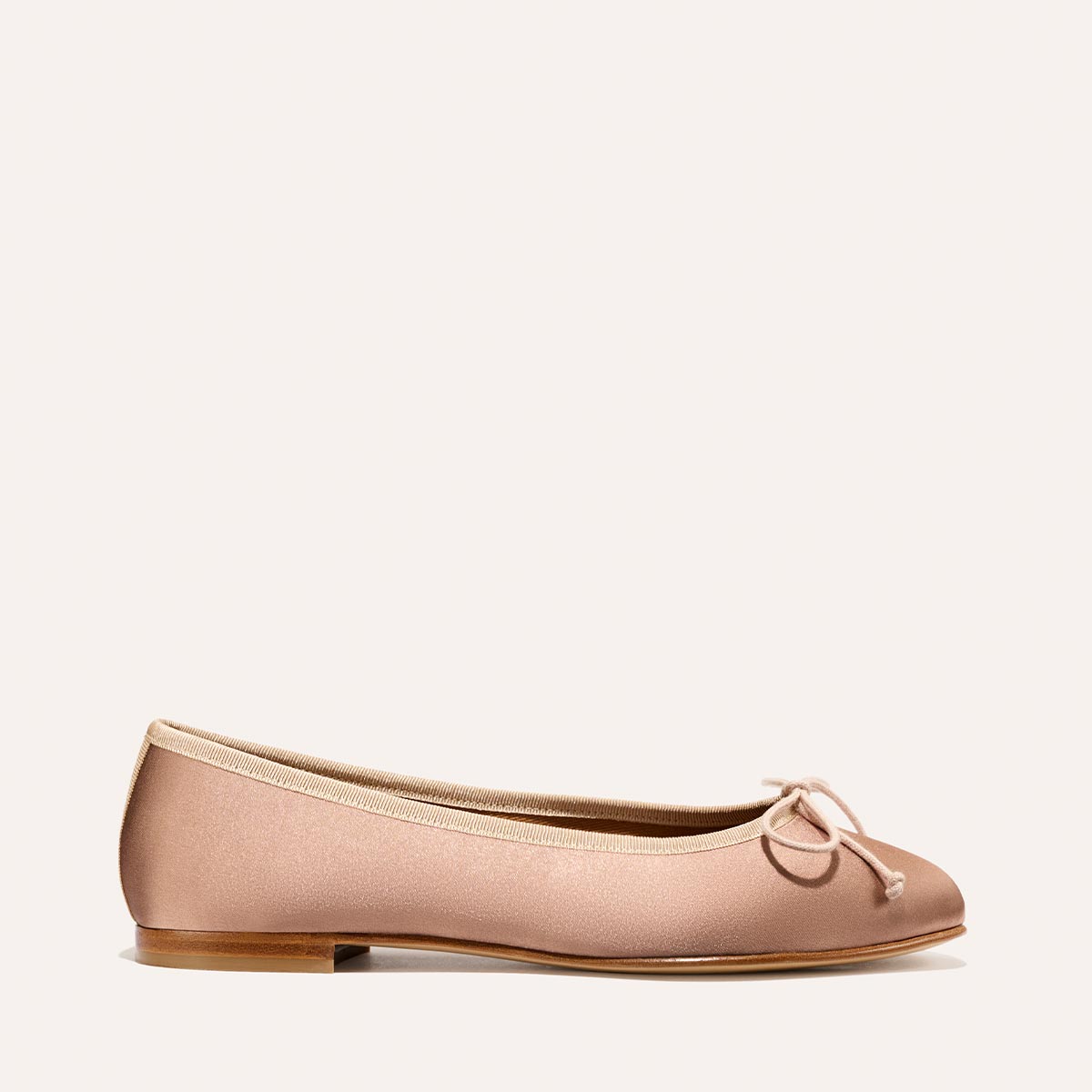 The Demi - Dusty Pink Satin by margaux-development is a handmade light pink ballet flat from Spain, featuring a small bow, beige trim, and a flat wooden sole, showcased on a simple light background.