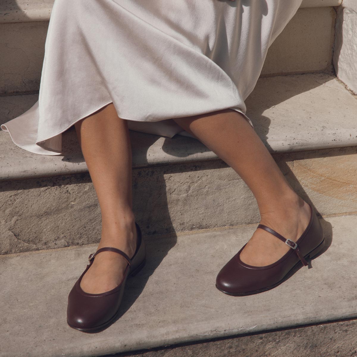 A person in a white dress sits on stone steps, exuding elegant femininity as they cross their legs and wear Margauxs The Demi Jane flats in Burgundy Nappa.