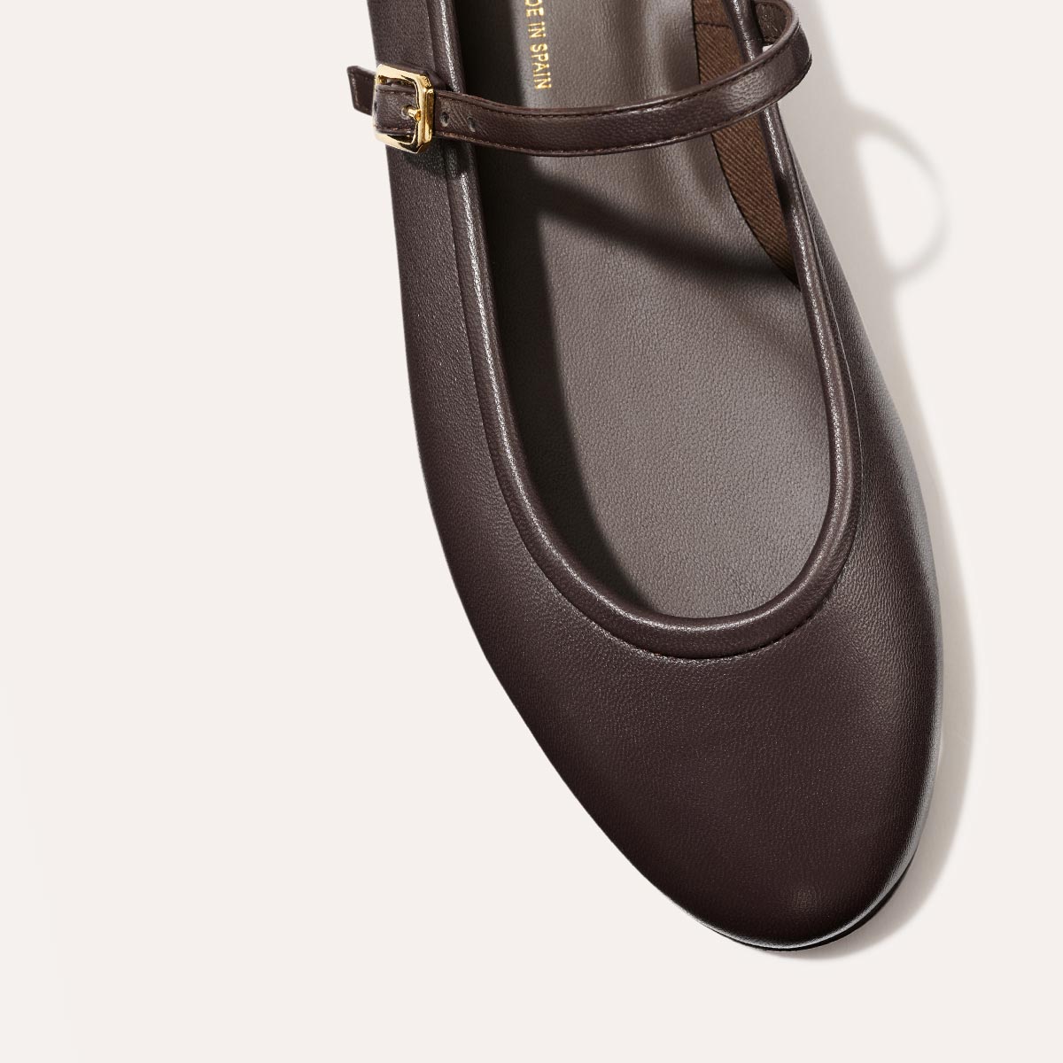 A close-up of The Demi Jane - Espresso Nappa by margaux-development, a dark brown leather flat with a thin strap and gold buckle, shown from above on a light background, showcasing elegant femininity.