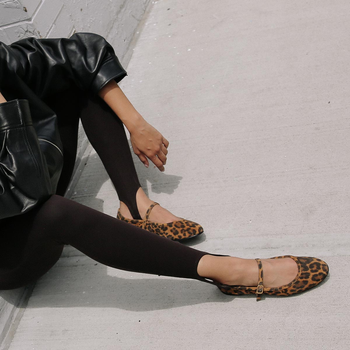 Exuding elegant femininity, she sits on a sidewalk in black leggings, a black leather jacket, and The Demi Jane - Leopard Suede flats by margaux-development, with one knee bent and her arm resting casually on her leg.