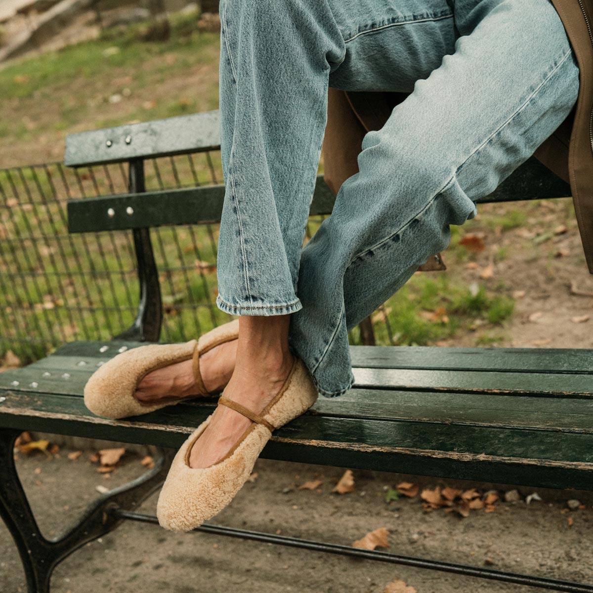 A person wearing light blue jeans and The Demi Jane - Tan Shearling by margaux-development sits elegantly on a green park bench, legs crossed, with fallen leaves and grass in the background.